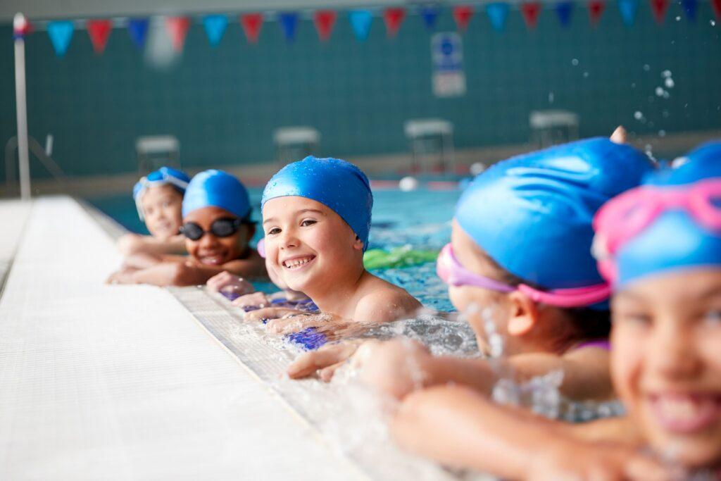 Kinder mit Schwimmkappen halten sich am Beckenrand eines Schwimmbades fest