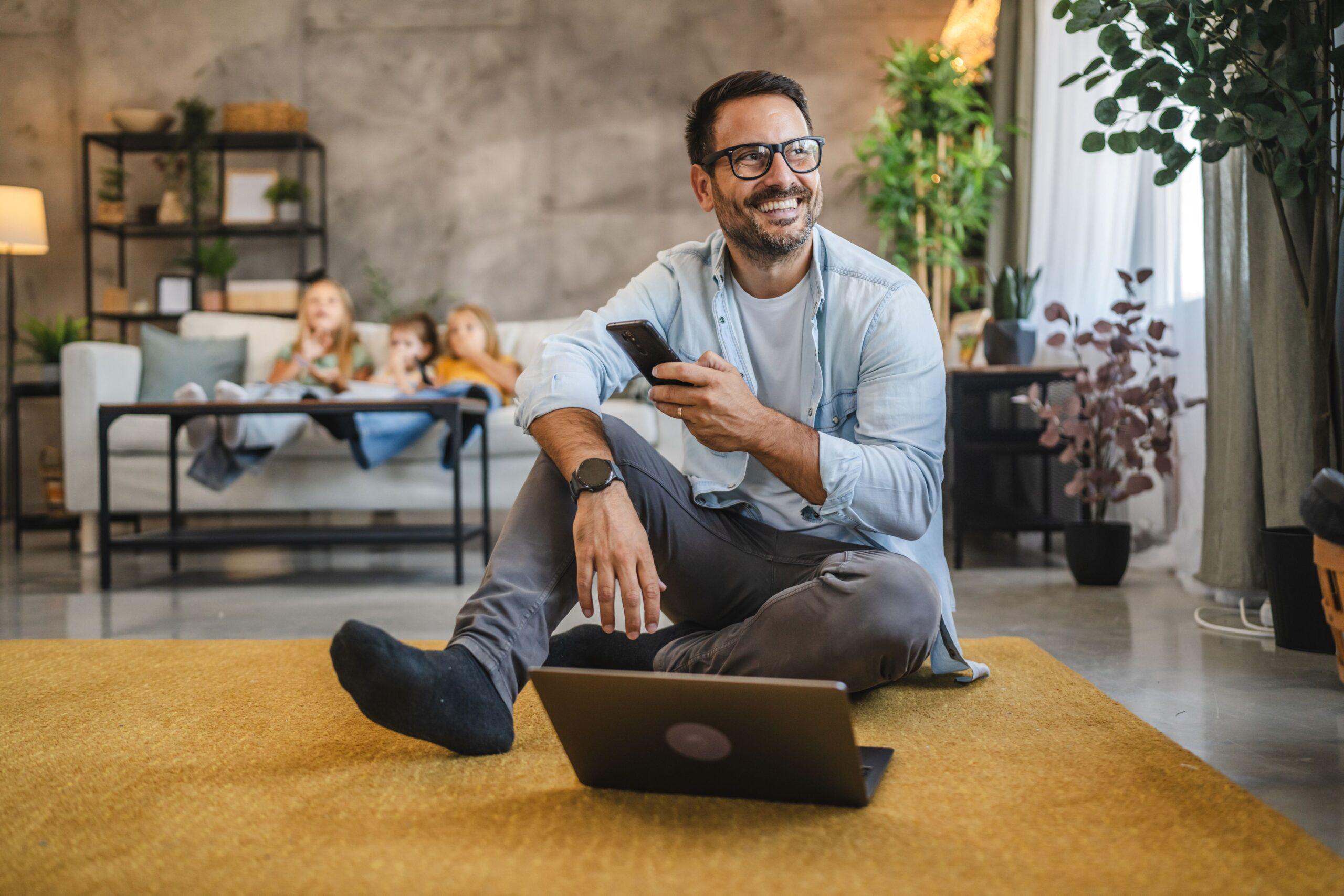 Ein Mann sitzt auf dem Boden vor einem Laptop hinter ihm sitzen Kinder auf einer Couch