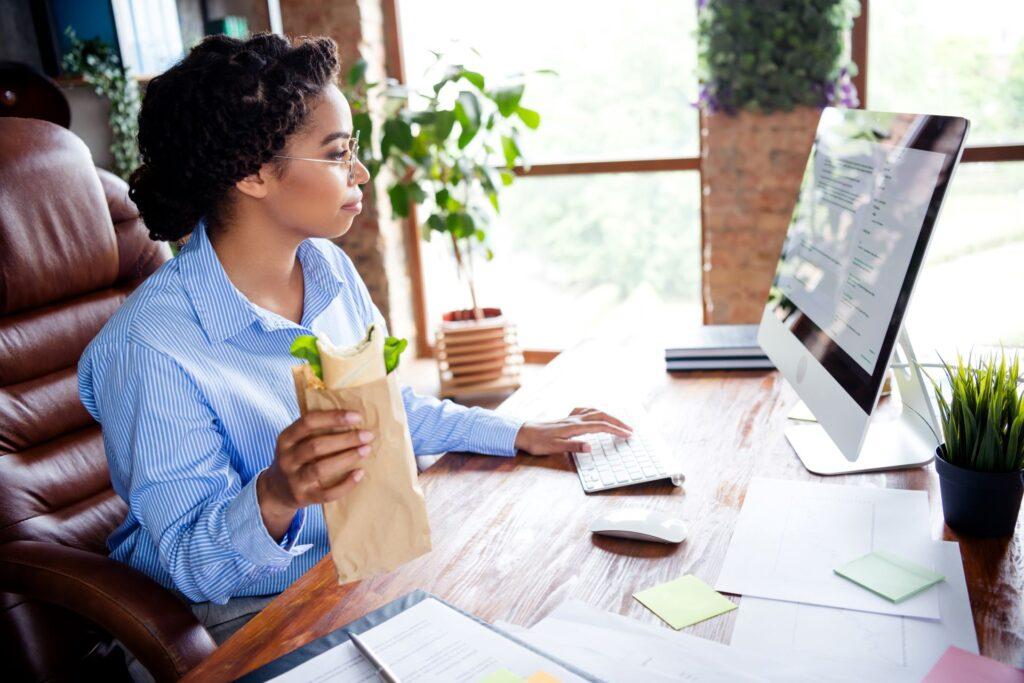 Frau am Schreibtisch mit Snack in der Hand, Work-Life-Balance im Fokus.